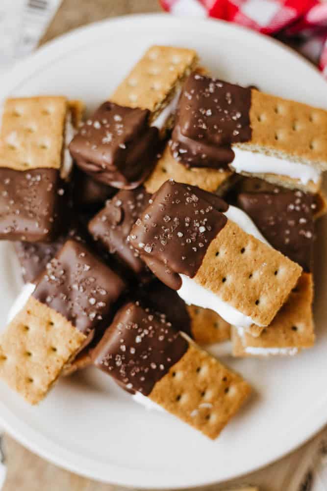 A plate of 3-ingredient S'mores Bites: rectangular graham cracker sandwiches filled with white marshmallow, partially dipped in chocolate, and sprinkled with coarse sea salt.