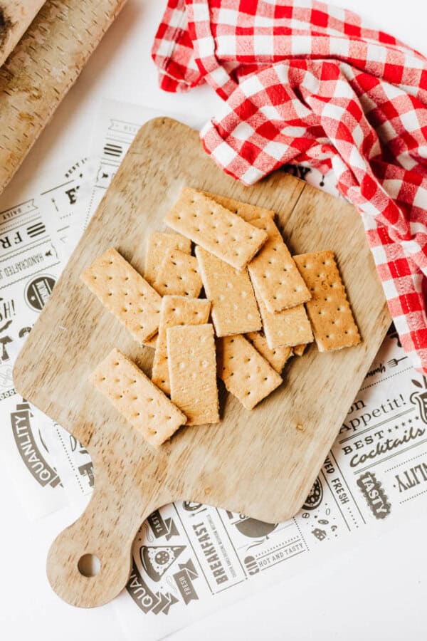 Rectangular crackers are arranged on a small wooden cutting board, next to a red and white checkered cloth atop food-themed decorative paper—perfect for serving 3-ingredient S'mores Bites.