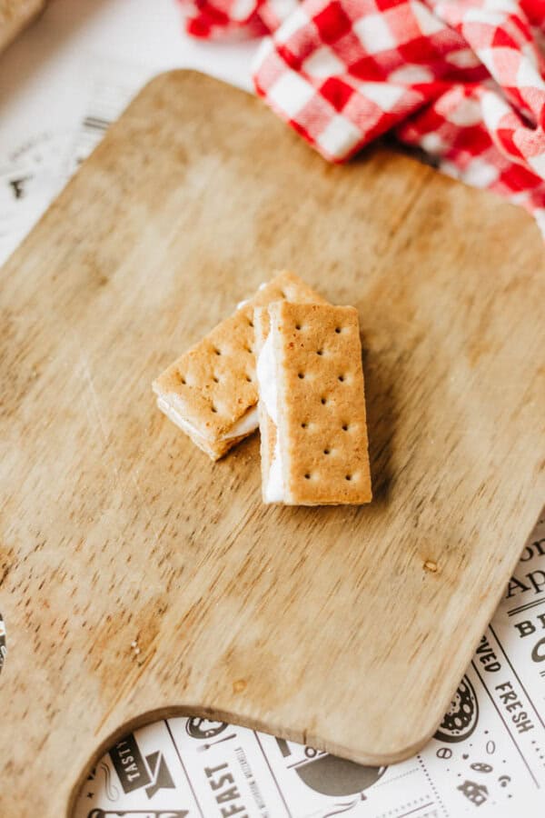 Two rectangular ice cream sandwiches on a wooden cutting board, with a red and white checkered cloth partially visible in the background—perfect alongside 3-ingredient S'mores Bites for an easy summer treat.