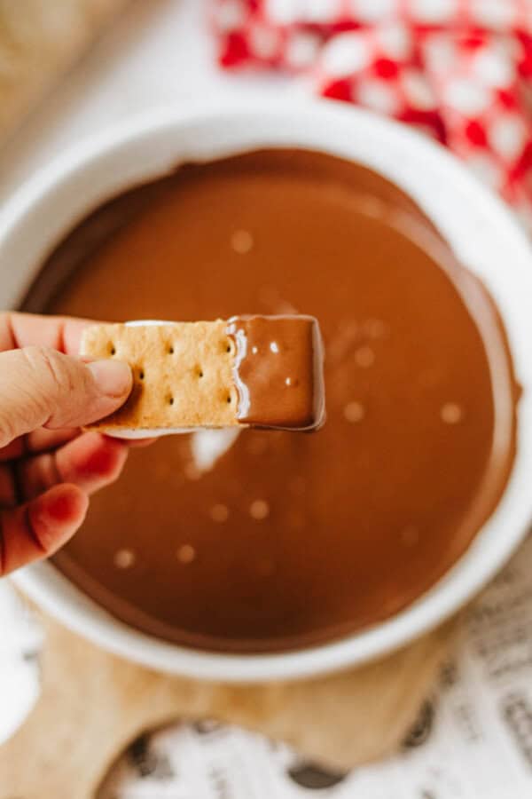 A hand holds a 3-ingredient S'mores Bites graham cracker partially dipped in melted chocolate, with a red and white cloth in the background.