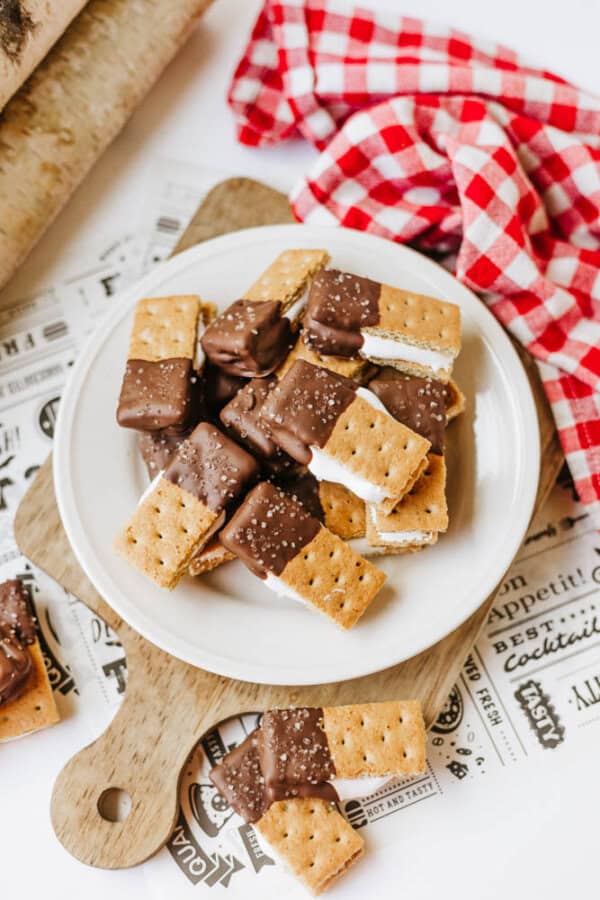 A plate of 3-ingredient S'mores Bites made with graham crackers, marshmallow, and chocolate are half-dipped in chocolate and sprinkled with salt. They sit on a round wooden board next to a red gingham cloth.