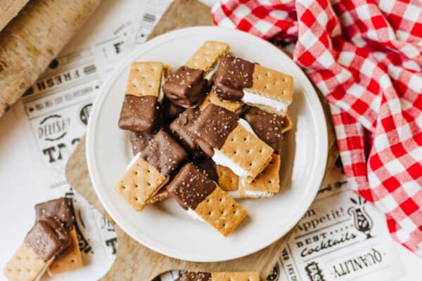 A plate of 3-ingredient S'mores Bites—ice cream sandwiches made with graham crackers and dipped in chocolate, sprinkled with salt—sits on a white plate next to a red and white checkered cloth.