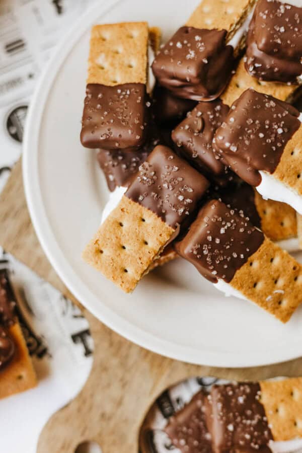 A plate of 3-ingredient S'mores Bites made with graham crackers, marshmallow, and chocolate—each half-dipped in chocolate and sprinkled with coarse sea salt. Some S'mores Bites rest on a wooden board nearby.