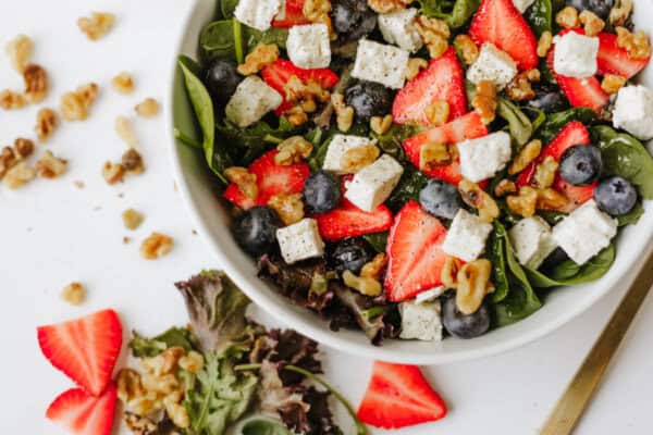 A fresh spinach strawberry and feta salad in a bowl with blueberries, walnuts, and cubes of feta cheese. Some salad ingredients and strawberry slices are scattered on the white surface nearby.