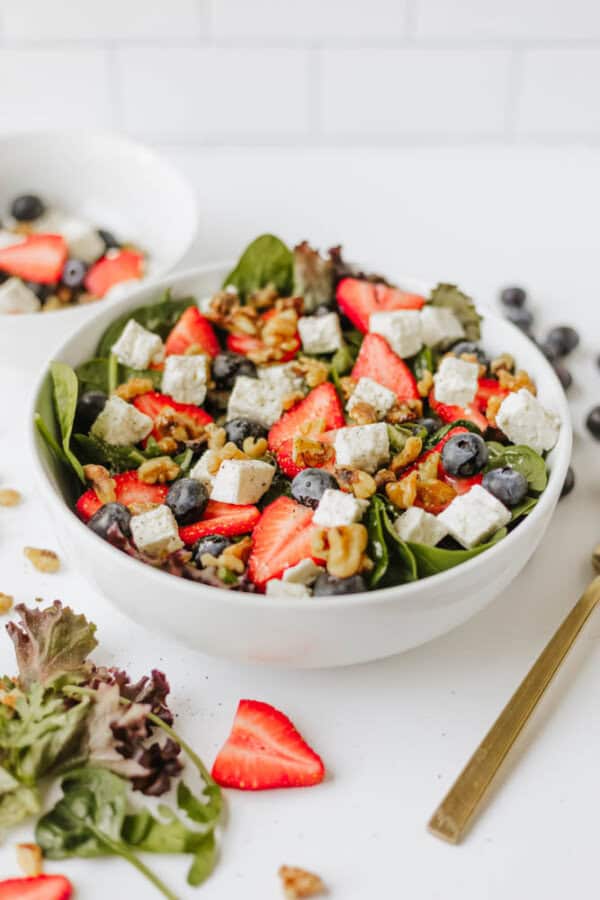 A white bowl filled with a vibrant spinach strawberry and feta salad, featuring blueberries, walnuts, and cubes of feta cheese, with a fork and extra salad greens nearby on a white surface.