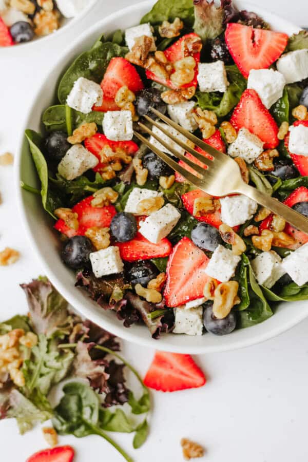 A bowl of fresh spinach strawberry and feta salad with blueberries, walnuts, and cubes of white cheese, topped with a gold fork. Some salad and fruit pieces are scattered on the white surface nearby.