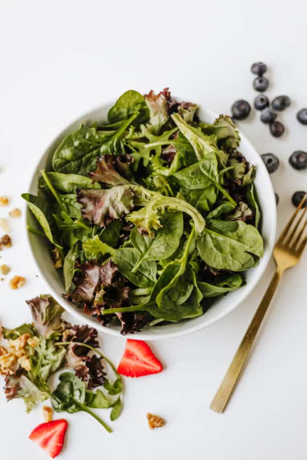 A bowl of mixed leafy greens on a white surface, featuring a vibrant spinach strawberry and feta salad with scattered blueberries, walnuts, and strawberry slices, plus a gold fork nearby.
