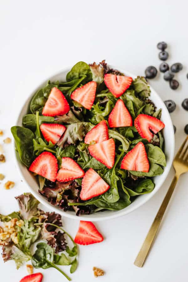 A white bowl filled with mixed green leafy spinach strawberry and feta salad, topped with fresh strawberry slices, surrounded by scattered blueberries, walnuts, and a gold fork on a white surface.