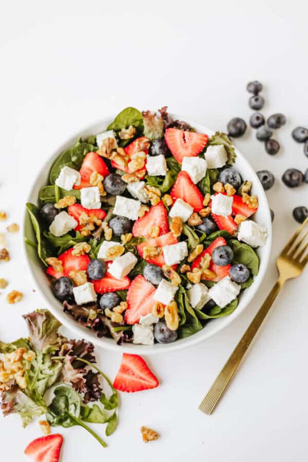 A bowl of spinach strawberry and feta salad features lettuce, blueberries, walnuts, and cubes of feta cheese. A gold fork and scattered berries and nuts rest beside the bowl on a white surface.