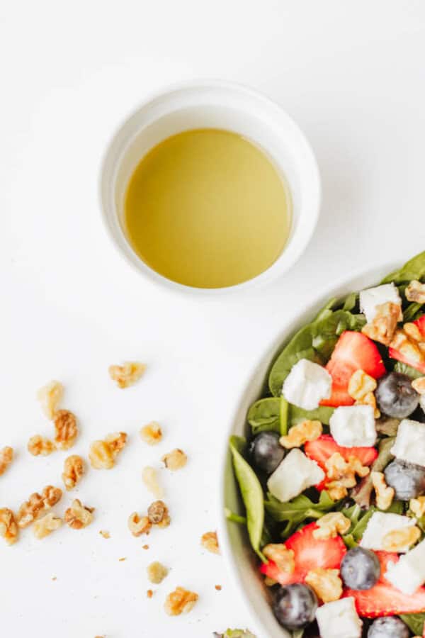 A bowl of spinach strawberry and feta salad with blueberries and walnuts sits next to a small white cup of olive oil, with extra walnuts scattered on the white surface.