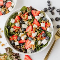 A bowl of spinach strawberry and feta salad features fresh spinach, strawberries, blueberries, feta cheese cubes, and walnuts. A gold fork, scattered berries, and salad ingredients are artfully arranged on a white surface nearby.