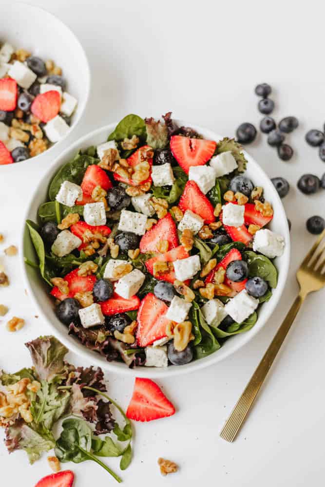 A bowl of spinach strawberry and feta salad features fresh spinach, strawberries, blueberries, feta cheese cubes, and walnuts. A gold fork, scattered berries, and salad ingredients are artfully arranged on a white surface nearby.