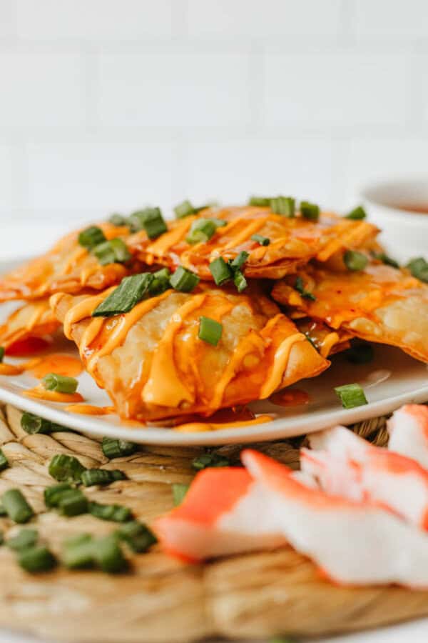 A plate of crispy, golden-brown Crab Rangoon Nachos drizzled with orange sauce and garnished with chopped green onions, with pieces of imitation crab in the foreground.