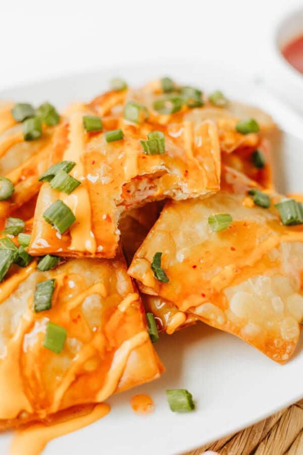 A close-up of crispy Crab Rangoon Nachos topped with orange sauce and chopped green onions, arranged on a white plate. One wonton has a bite taken out, revealing the creamy filling inside.