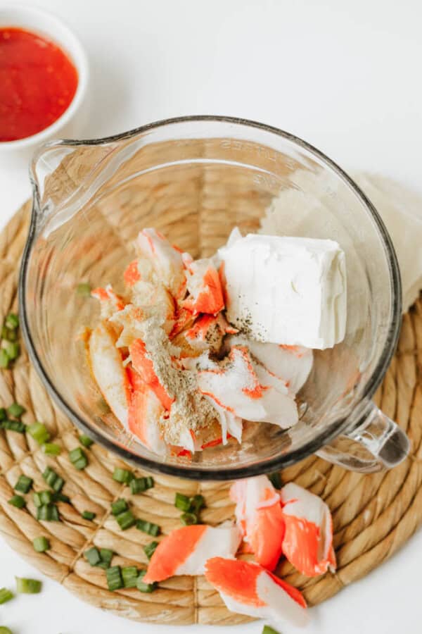 A glass mixing bowl with imitation crab sticks, a block of cream cheese, and seasonings sits on a woven mat—perfect for making Crab Rangoon Nachos—with chopped green onions and a small bowl of red sauce nearby.