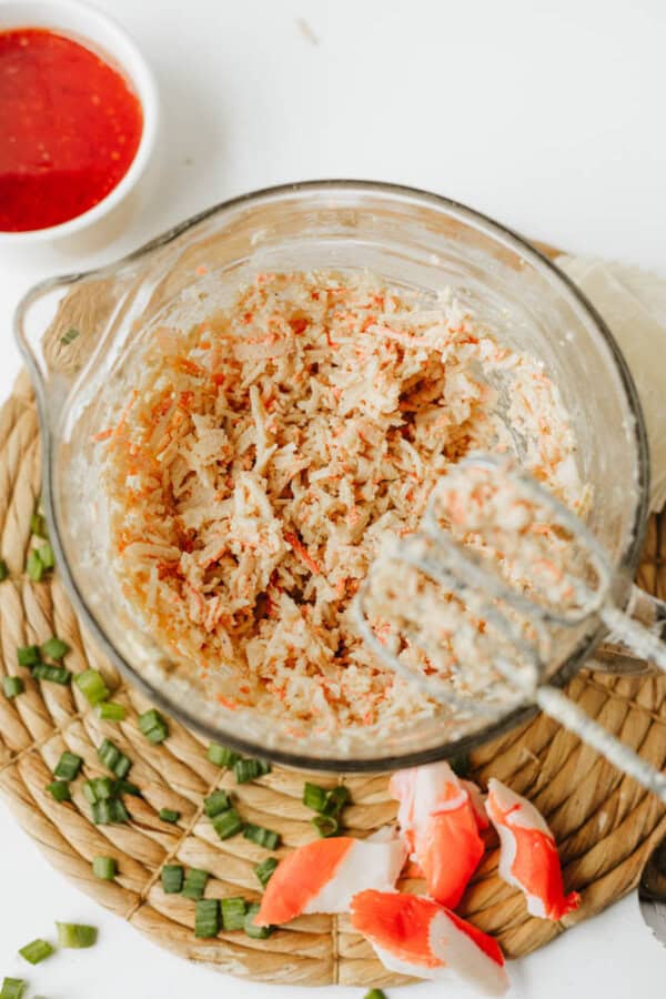 A glass mixing bowl with shredded crab mixture for Crab Rangoon Nachos sits on a woven mat, surrounded by crab sticks, chopped green onions, and a small bowl of red dipping sauce. An electric mixer is partially visible in the bowl.