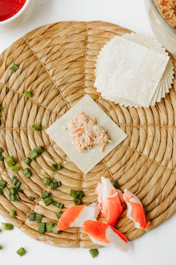 A square wonton wrapper with crab filling sits on a woven mat, surrounded by green onion pieces, imitation crab sticks, a stack of wonton wrappers, and a bowl of sauce—perfect ingredients for Crab Rangoon Nachos.