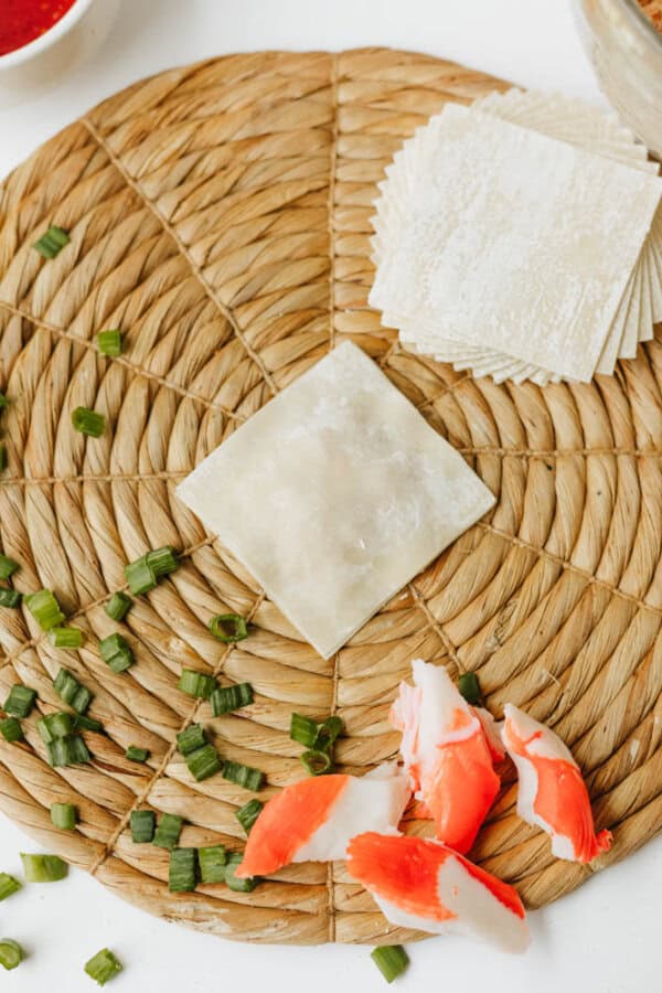 Square wonton wrappers and a filled wonton sit on a woven placemat, surrounded by chopped green onions and imitation crab meat pieces—perfect ingredients for homemade Crab Rangoon Nachos.