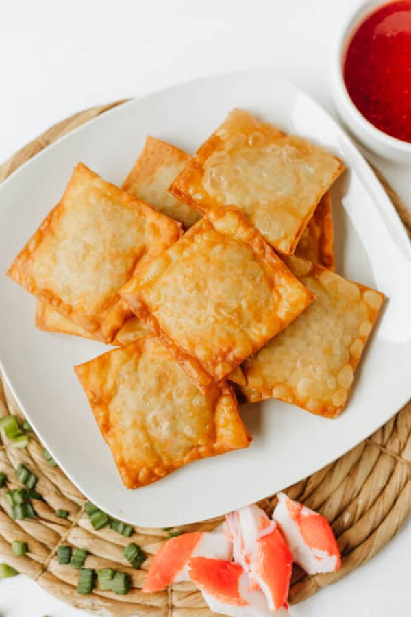 A white plate with six golden-brown Crab Rangoon Nachos sits on a woven placemat, next to chopped green onions, imitation crab pieces, and a small bowl of red dipping sauce.