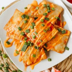 A white plate of crispy Crab Rangoon Nachos drizzled with orange sauce and garnished with chopped green onions, served on a woven mat with imitation crab and a bowl of dipping sauce nearby.