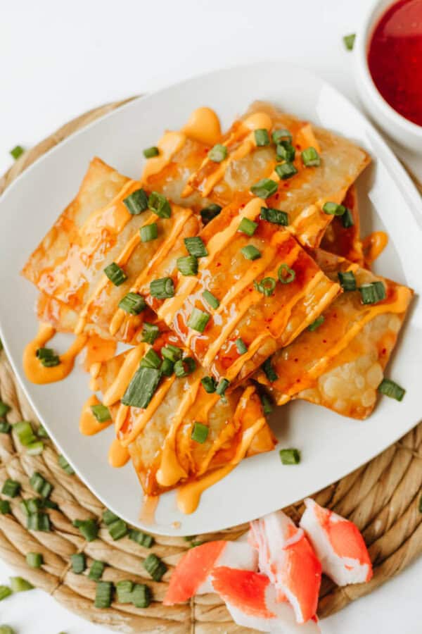 A white plate of crispy Crab Rangoon Nachos drizzled with orange sauce and garnished with chopped green onions, served on a woven mat with imitation crab and a bowl of dipping sauce nearby.