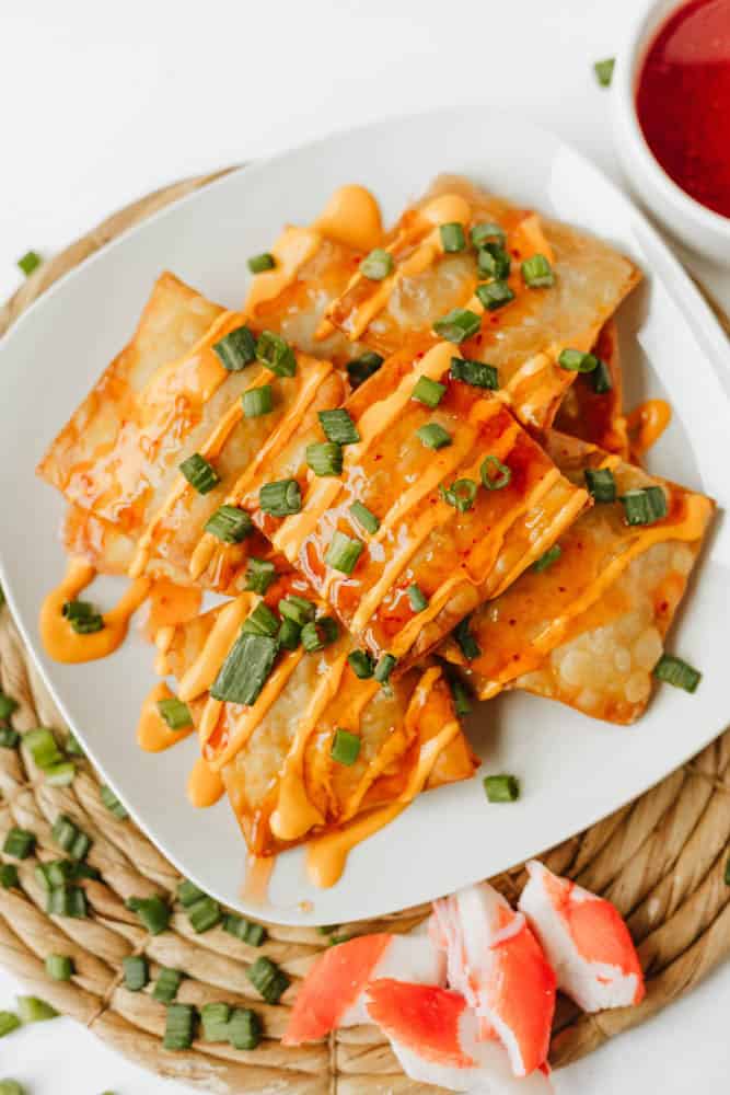 A white plate of crispy Crab Rangoon Nachos drizzled with orange sauce and garnished with chopped green onions, served on a woven mat with imitation crab and a bowl of dipping sauce nearby.