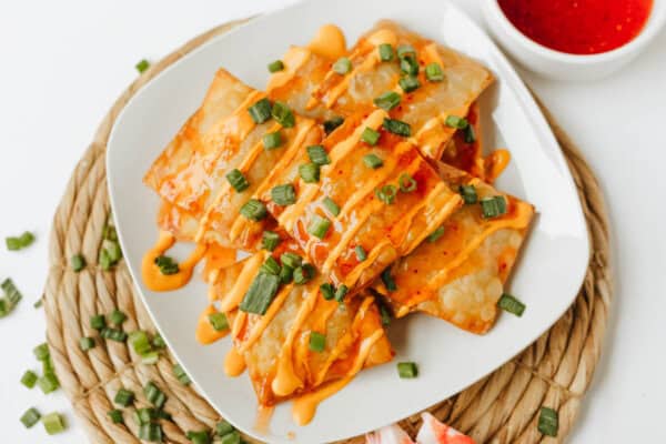 A white plate with crispy Crab Rangoon Nachos drizzled with orange sauce and sprinkled with chopped green onions, placed on a woven placemat with a small bowl of red dipping sauce beside it.