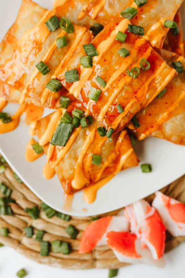 A plate of crispy wontons reminiscent of Crab Rangoon Nachos, drizzled with orange sauce and topped with chopped green onions, sits on a woven mat with imitation crab pieces and scattered green onions nearby.