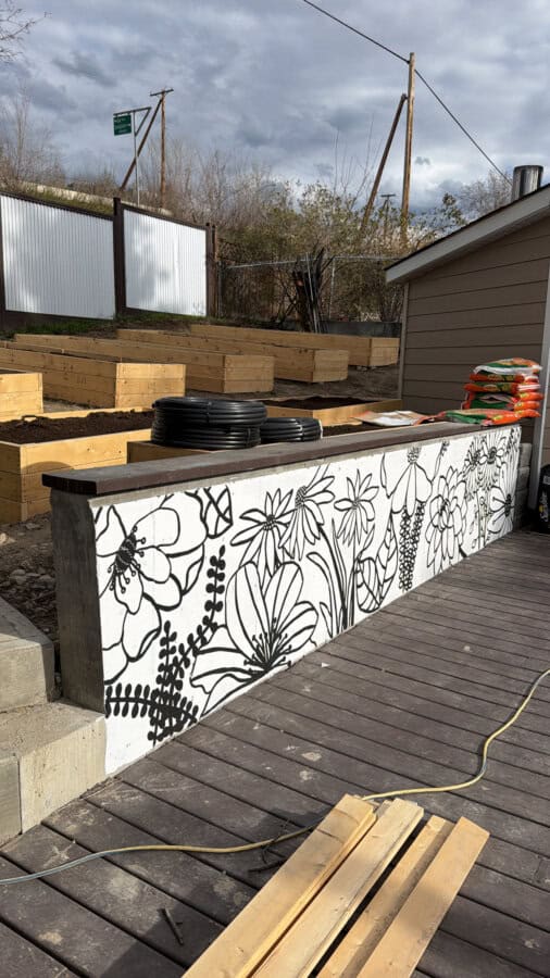 A raised garden bed with a black and white floral mural sits on a wooden deck. Stacks of pots and bags of soil are nearby, with tiered beds and trees in the background under a cloudy sky.