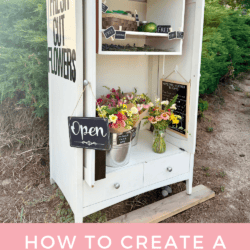 A white flower stand with shelves holds fresh cut flowers, chalkboard signs, and a metal bucket of blooms. A large sign reads FRESH CUT FLOWERS. The stand sits outdoors with greenery in the background.