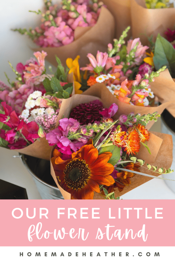 Bouquets of colorful fresh flowers wrapped in brown paper are displayed in a metal bucket. A pink sign below reads, Our Free Little Flower Stand homemadheather.com.
