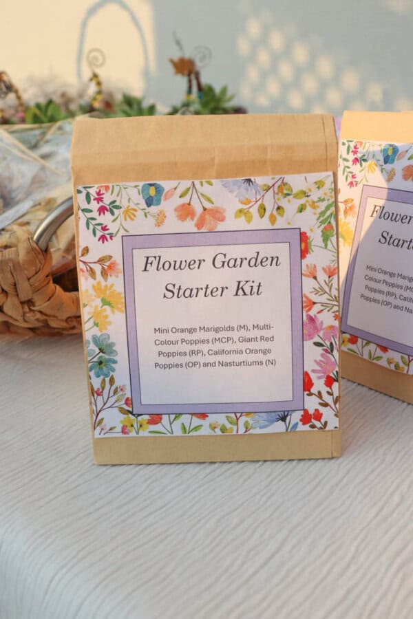A paper bag labeled Flower Garden Starter Kit with a floral border, listing marigold, poppy, and nasturtium seeds. The bag sits on a white cloth near a wicker basket.