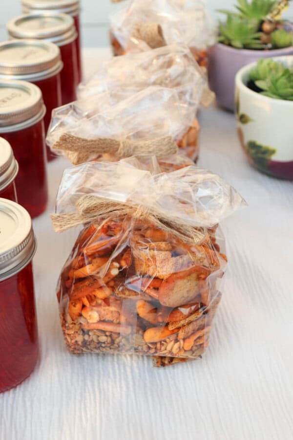 Clear gift bags filled with a snack mix are tied with burlap bows and lined up beside jars of red preserves. Succulent plants in decorative pots are visible in the background on a white tablecloth.