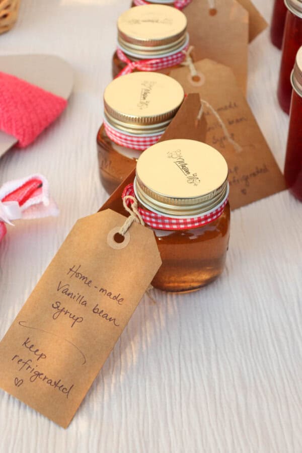 Three small glass jars with gold lids and gingham ribbons, labeled “Homemade Vanilla Bean Syrup.” Each jar has a brown gift tag with handwritten storage instructions. The jars are placed on a light textured surface.