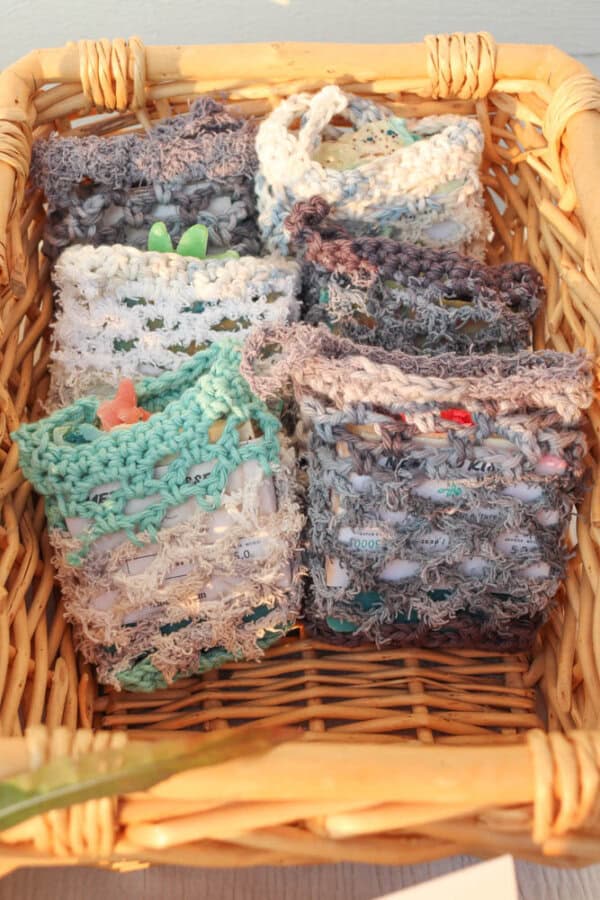 Wicker basket containing several small, handmade crochet pouches in various pastel colors, each filled with small objects and displayed neatly.