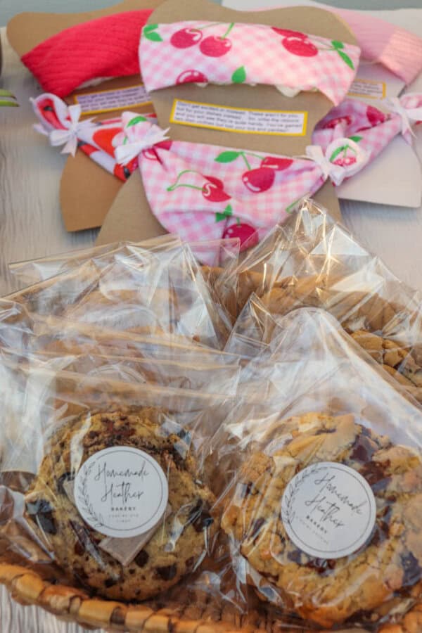 Individually wrapped cookies in a basket labeled “Homemade Heather Bakery,” with pink cherry-patterned cloth masks displayed in the background on a table.
