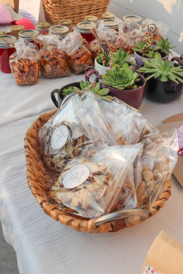 A basket of bagged cookies sits on a table next to jars of preserves, bags of snacks, and potted succulents in teacups, all arranged for a market or sale.