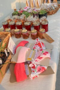A table displays jars of jam, bags of snacks, potted succulents, and handmade headbands in pink and red with tag labels, organized neatly at an outdoor market.