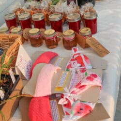 A table displays jars of jam, bags of snacks, potted succulents, and handmade headbands in pink and red with tag labels, organized neatly at an outdoor market.