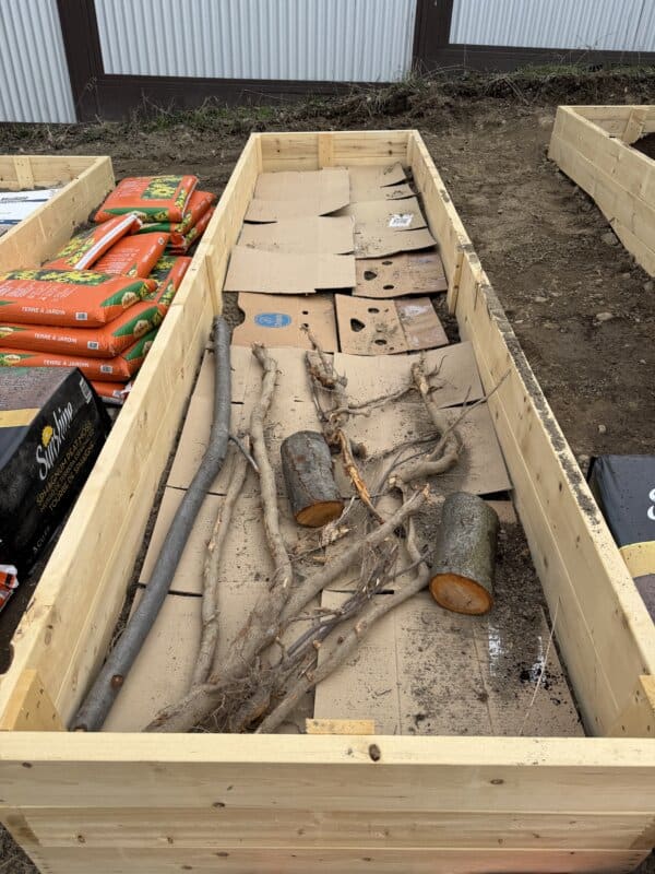 A wooden raised garden bed filled with cardboard, branches, and logs, surrounded by bags of soil and mulch. The ground is bare soil, and a corrugated metal fence is in the background.