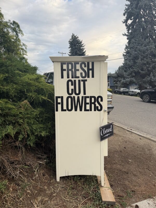 A white cabinet with large black letters reading FRESH CUT FLOWERS stands outside; a small sign hanging from the handle says Closed. Shrubs, trees, cars, and a street are visible in the background.