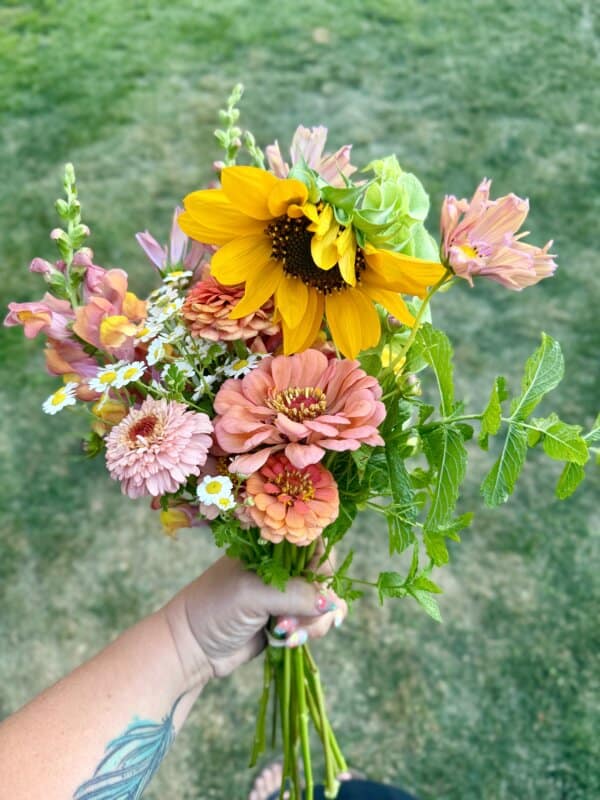 A hand holding a colorful bouquet of flowers, including sunflowers, zinnias, daisies, and greenery, against a blurred grassy background. The person has a tattoo on their forearm.