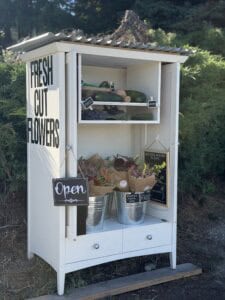 A white outdoor stand with a sign reading Fresh Cut Flowers displays bouquets in metal buckets and some vegetables. An Open sign hangs on the door, and a Free label is on the veggies. Shrubs fill the background.
