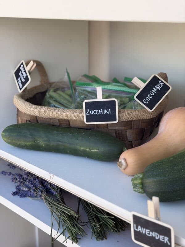 A basket with labeled bags of zucchini and cucumbers sits on a shelf next to whole zucchinis, cucumbers, a butternut squash, and a bundle of lavender, each labeled with small chalkboard signs.