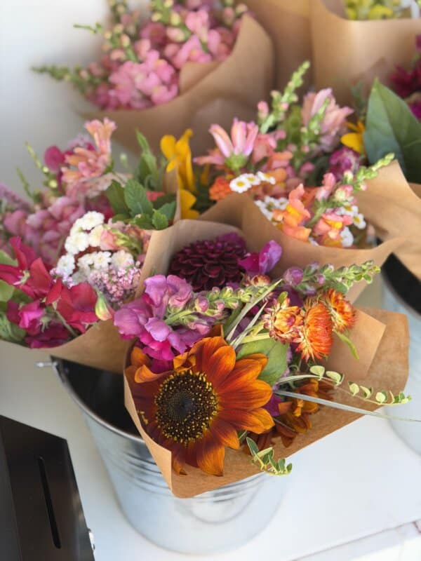 Several bouquets of colorful flowers, including sunflowers, snapdragons, and daisies, are wrapped in brown paper and displayed in a metal bucket. The bright, fresh flowers create a vibrant, cheerful arrangement.