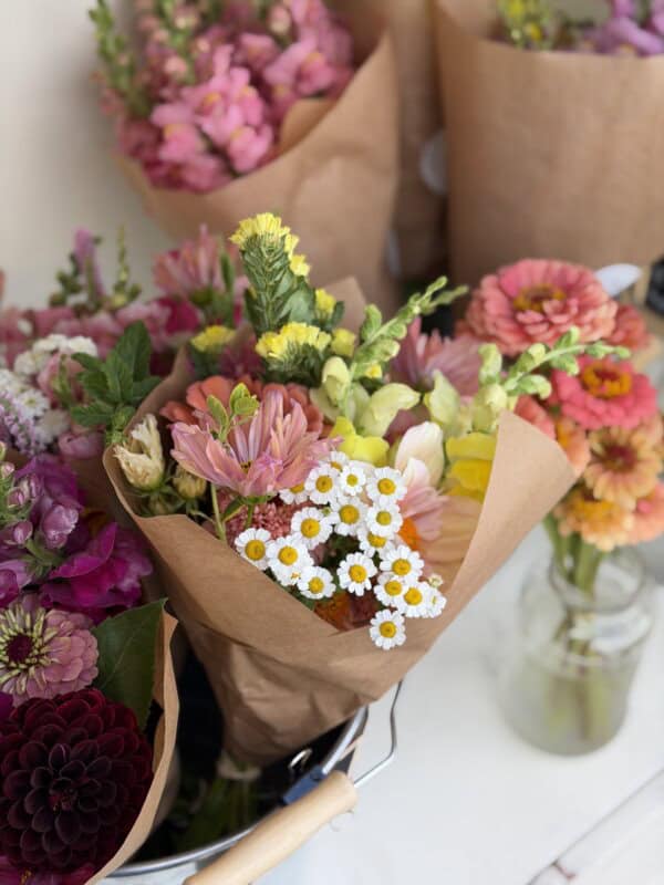 A bouquet of mixed flowers, including daisies, zinnias, and pink blossoms, wrapped in brown paper. Other bouquets and a glass jar with flowers are nearby on a white surface.