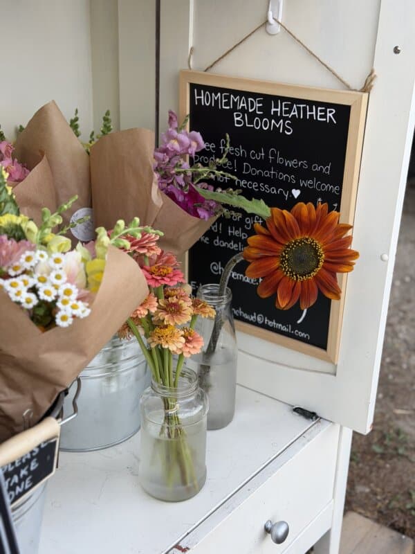 Bouquets of fresh flowers in jars and brown paper sit beside a chalkboard sign reading โHomemade Heather Blooms,โ with a large orange flower attached to the sign.
