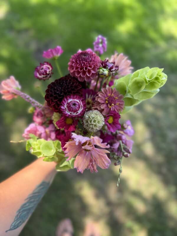 A hand with a tattoo holds a colorful bouquet of pink, purple, and green flowers against a blurred grassy background.