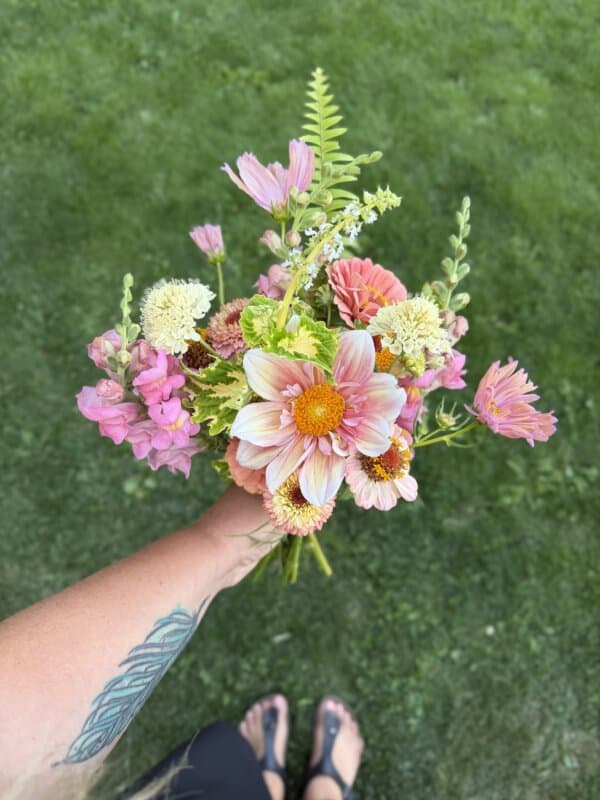 A hand holding a colorful bouquet of flowers with pink, yellow, and orange blooms, above green grass. A leaf tattoo is visible on the forearm, and feet in sandals appear at the bottom of the image.