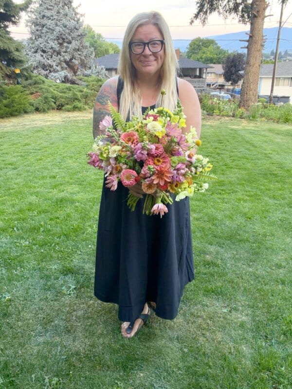 A woman with long blonde hair and glasses stands on grass holding a large bouquet of colorful flowers. She wears a sleeveless black dress and sandals, and there are trees and houses in the background.